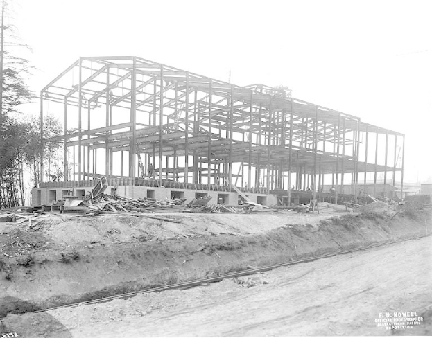 Ein Schwarz-Weiß-Foto eines Gebäudes im Bau mit einer durchlaufenden Bahnstrecke, Bäumen links und einem klaren Himmel im Hintergrund, mit Text unten, der es als Foto des Neubaus einer Schule identifiziert.