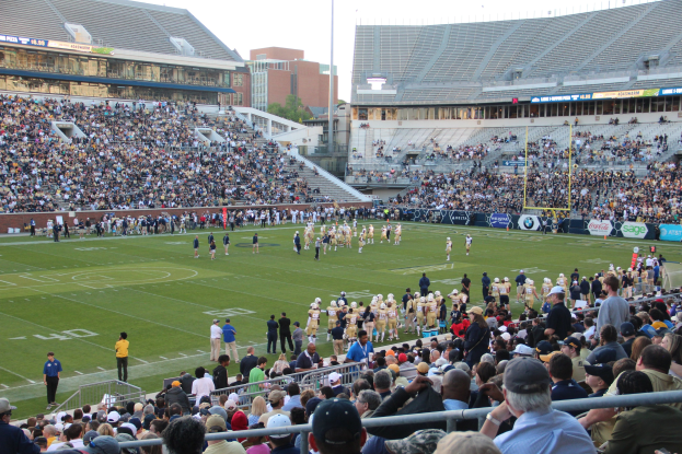 Ein Fußballspiel im Gange mit Spielern auf dem Feld und Zuschauern in den Tribünen, in einem Stadion unter einem klaren blauen Himmel.