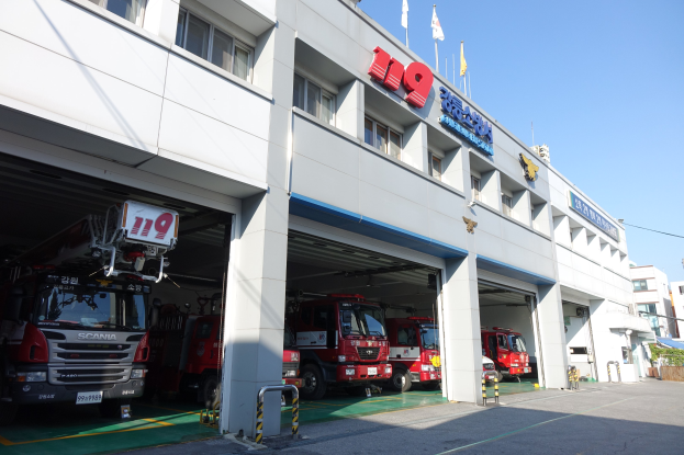 Feuerwache mit mehreren Feuerwehrautos vor der Tür, mit Fenstern, Säulen, Namensschildern und Flaggen im Vordergrund vor einem Hintergrund aus Gebäuden und einem klaren blauen Himmel.