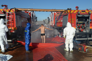 Drei Personen in hochwertiger Jacke stehen im Vordergrund, mit einer Person auf einer Abdeckung mit Wasser davor. Several others and fire engine trucks with extended ladders are in the middle ground, and buildings are visible in the background.