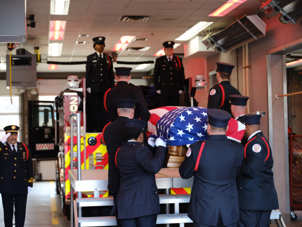 Gruppe uniformierter Männer, die einen Sarg mit einer amerikanischen Flagge tragen, umgeben von Menschen mit Mützen und einem Löschfahrzeug sowie Deckenbeleuchtung im Hintergrund.