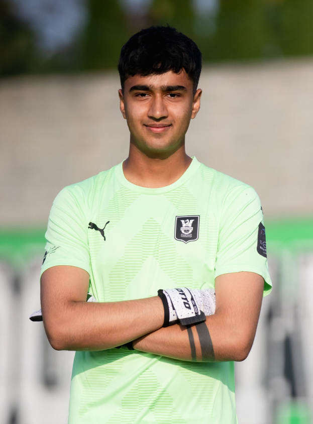 Ein junger Mann in einem grünen Hemd steht auf einem Fußballfeld und schaut mit verschränkten Armen und einem Lächeln in die Kamera, mit einem unscharfen Hintergrund.