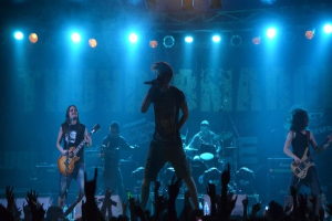 Eine Gruppe von Musikern auf einer hell erleuchteten Bühne, einige spielen Gitarren, einer singt in ein Mikrofon und ein anderer spielt Schlagzeug im Hintergrund, während Zuschauer im Vordergrund die Hände heben.