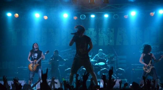 Eine Gruppe von Musikern auf einer hell erleuchteten Bühne, einige spielen Gitarren, einer singt in ein Mikrofon und ein anderer spielt Schlagzeug im Hintergrund, während Zuschauer im Vordergrund die Hände heben.