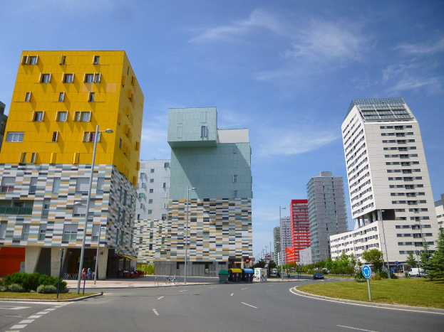 Eine Stadtstraße mit hohen Gebäuden im Hintergrund, Straßenlaternen, Autos, Informationsschildern, Büschen, Bäumen und einem bewölkten Himmel, wobei die Gebäude der Universität Rotterdam gehören.