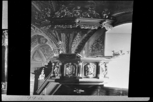 Ein Schwarz-Weiß-Foto eines Kirchenpults mit geschnitzten Säulen und einer strukturierten Wand im Hintergrund, das unten einen Text zeigt.