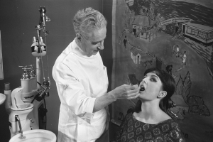 Ein Schwarz-Weiß-Bild eines Zahnarztes, der eine sitzende Frau untersucht, mit einem Waschbecken links und verschiedenen Gegenständen rechts und einem Gemälde im Hintergrund.