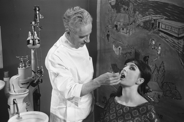 Ein Schwarz-Weiß-Bild eines Zahnarztes, der eine sitzende Frau untersucht, mit einem Waschbecken links und verschiedenen Gegenständen rechts und einem Gemälde im Hintergrund.