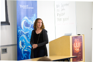 Eine Frau in einem schwarzen Kleid steht neben einem Rednerpult in einem Konferenzraum, mit einem Tisch daneben und einem Banner, einer Wand und einem Fenster im Hintergrund.