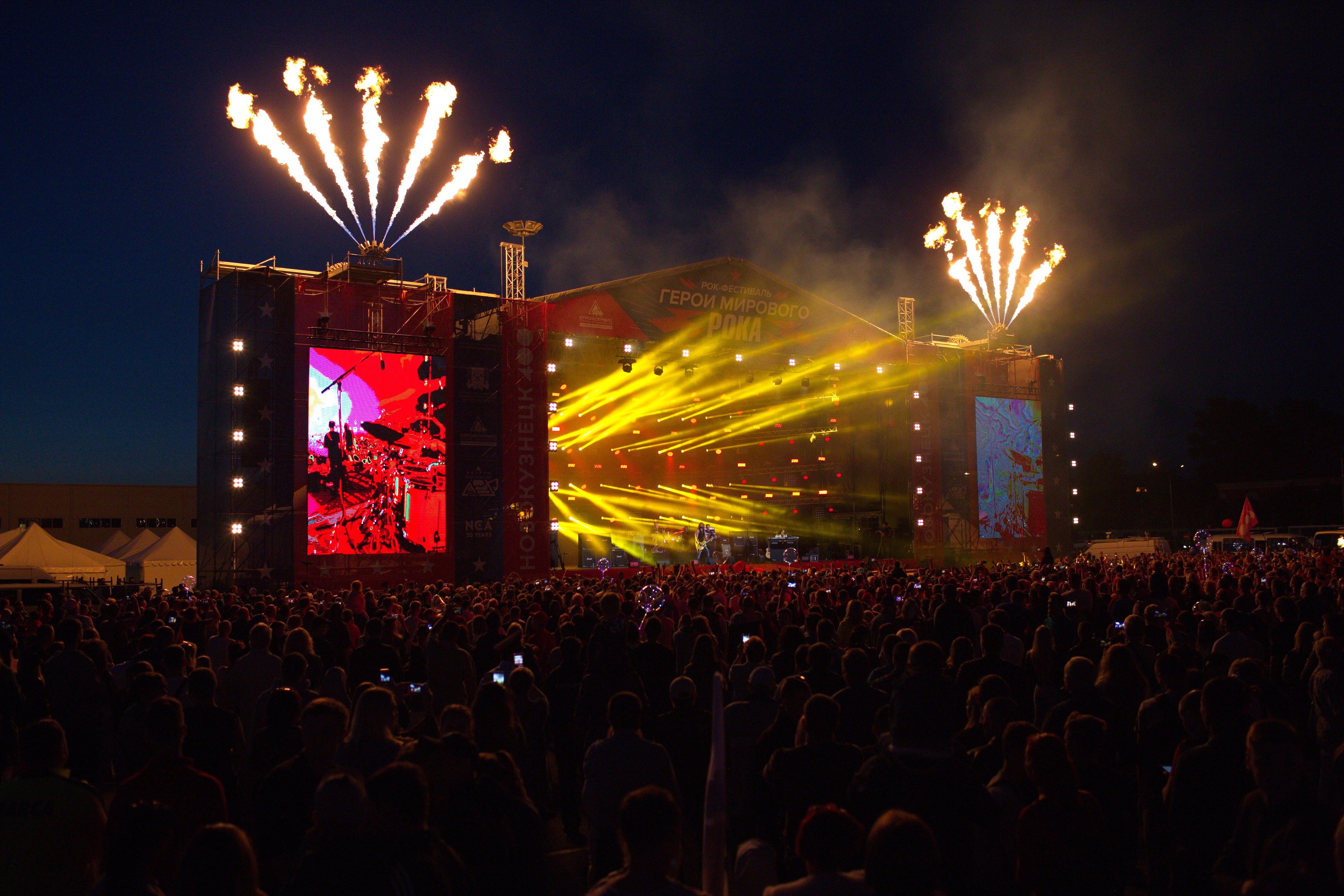 Eine große Menschenmenge auf einem Musikfestival schaut auf die Bühne mit hellen Lichtern, links Zelte und ein dunkler Himmel mit Feuerwerk.