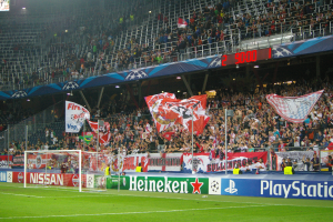 Ein Fußballspiel wird in einem gut beleuchteten Stadion mit einer großen Menge, grünem Rasen, einem Torpfosten mit Bannern und einem Bildschirm in der oberen linken Ecke gespielt.