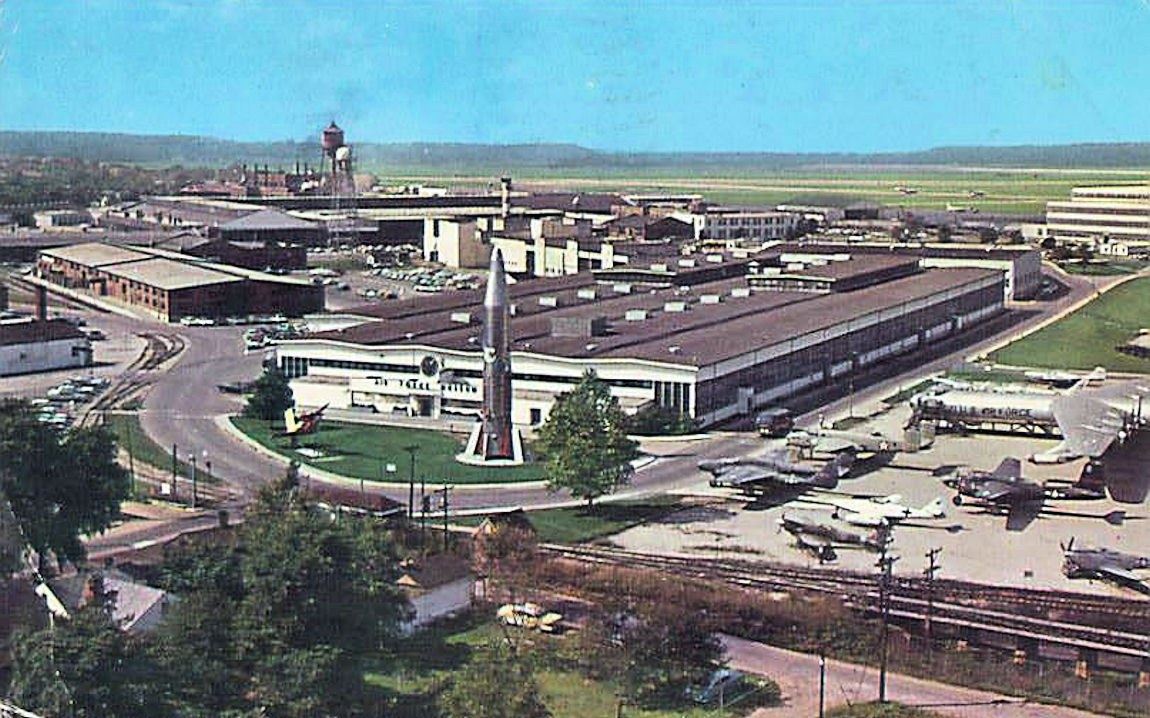 Altes Schwarz-Weiß-Foto eines Flughafens mit parkenden Flugzeugen, Gebäuden, Bäumen, Straßen, Fahrzeugen, Strommasten, Gras, Bahnschienen, Türmen, Hügeln und einem klaren Himmel, mit Text entlang der unteren Kante.