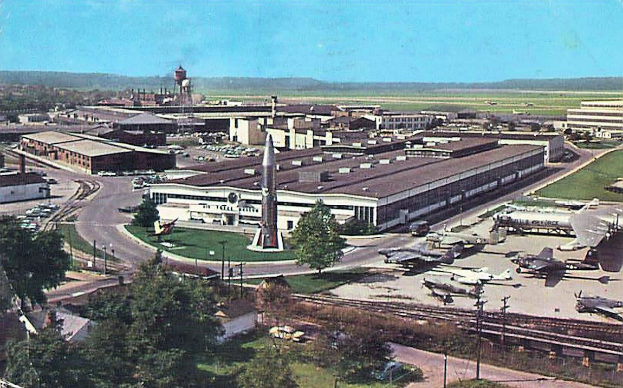 Altes Schwarz-Weiß-Foto eines Flughafens mit parkenden Flugzeugen, Gebäuden, Bäumen, Straßen, Fahrzeugen, Strommasten, Gras, Bahnschienen, Türmen, Hügeln und einem klaren Himmel, mit Text entlang der unteren Kante.