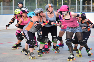 Mehrere Frauen mit Helmen und Schlittschuhen schieben sich gegenseitig auf Kufen, mit einem Mann und einem Metallzaun im Hintergrund und Bäumen weiter hinten.