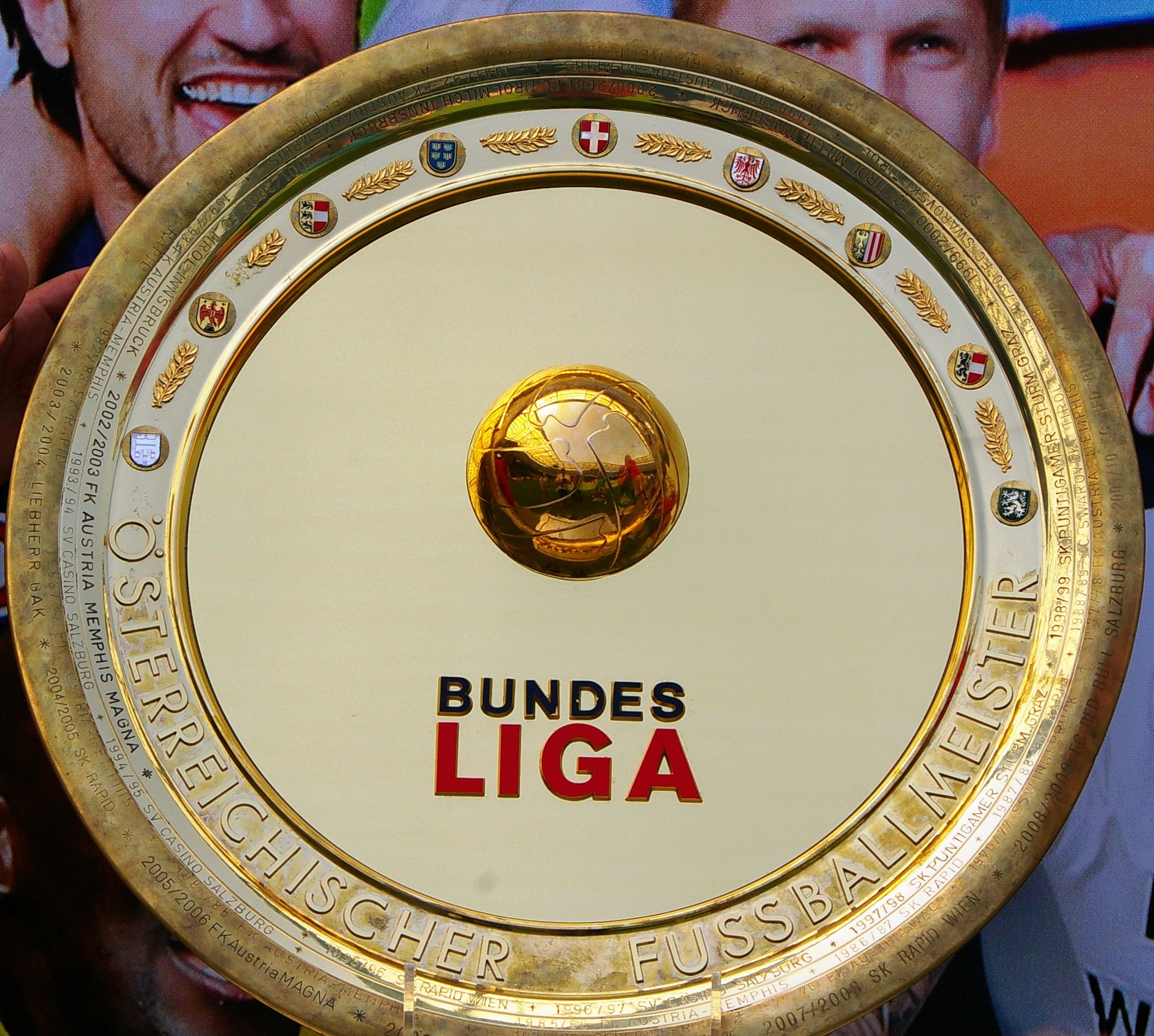 Ein Mann hält einen goldenen Schild mit der Aufschrift 'Bundesliga', im Hintergrund eine Fahne mit drei Personen.