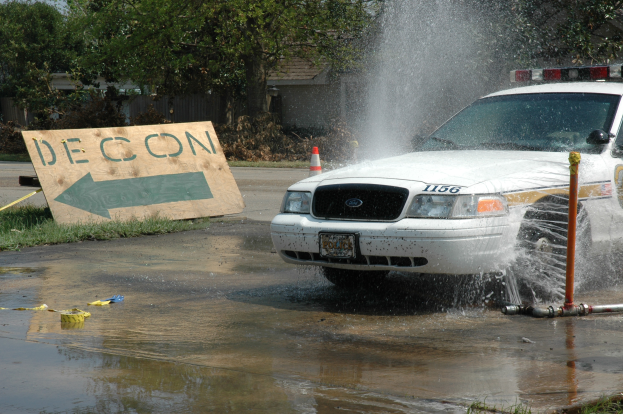 Ein Polizeiwagen fährt durch eine Pfütze, mit einem Schild, einem Verkehrskegel und anderen Gegenständen am Boden, mit Bäumen, Gebäuden und einem klaren blauen Himmel im Hintergrund.