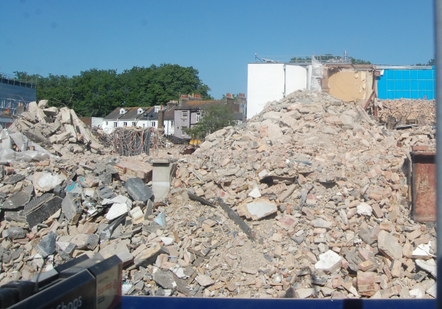 Großer Schutthaufen auf einer Baustelle mit einem Abrissgerät unten, umgeben von Gebäuden, Bäumen und einem klaren blauen Himmel.