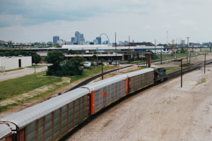 Güterzug fährt auf Schienen neben einer Stadtlandschaft mit Strommasten, Kabeln, Gebäuden, Bäumen, Gras und einem bewölkten Himmel.