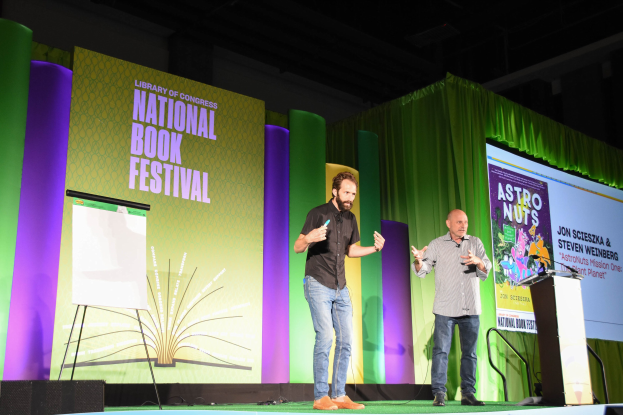 Zwei Männer stehen auf einer Bühne beim Nationalen Buchfestival, mit einem Podium und Mikrofon rechts und einem großen Bildschirm dahinter.
