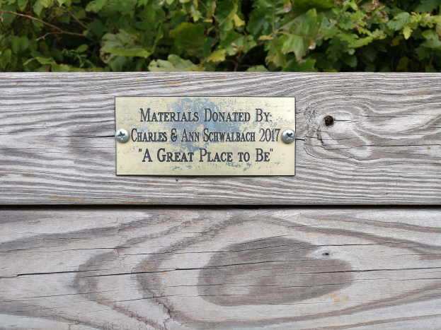 Holzbank mit einer Plakette, auf der "Materialien gespendet von Charles & Ann Schwabach 2017: Ein schöner Ort" steht, vor grünen Pflanzen.