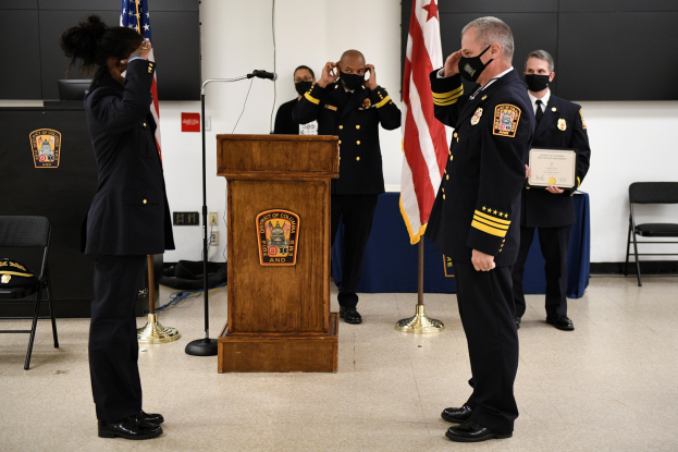 Gruppe uniformierter Personen in Masken vor einem Podium während einer Vereidigung eines neuen Feuerwehrchefs, der ein Zertifikat hält, mit Flaggen, Stühlen, Tischen und einer Wand im Hintergrund.