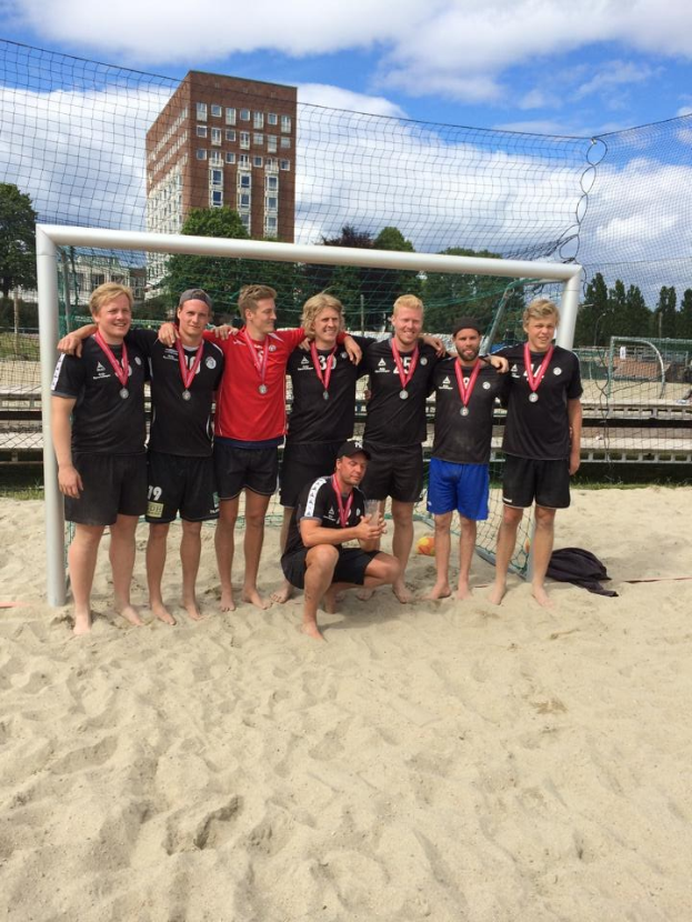 Mannergruppe in Sportkleidung auf sandigem Strand mit Medaillen, umgeben von einem Netz, mit Bäumen, Gebäuden und bewölktem Himmel im Hintergrund.