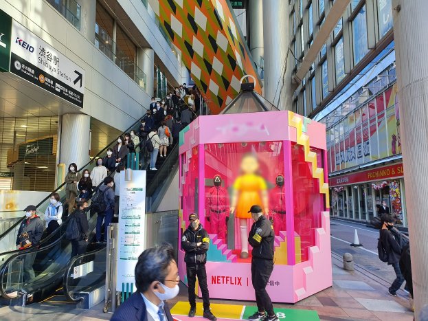 Eine Gruppe von Menschen steht vor einem Gebäude mit Glaswänden, Säulen und Texttafeln, einige tragen Masken, während eine Rolltreppe mit Menschen im Hintergrund zu sehen ist; ein pinkfarbener Karton mit Schaufensterpuppen darin, beschriftet mit 'Netflix' neuem Netflix-Stand in Tokyo', ist prominent zu sehen.