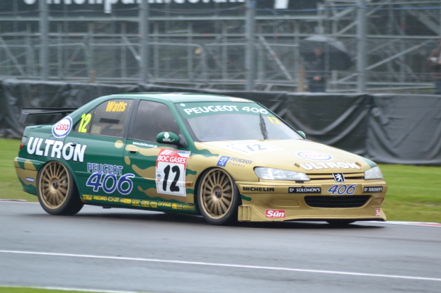 Ein Auto fährt auf einer Rennstrecke mit Gras an beiden Seiten, ein Rad im Vordergrund und Zuschauern mit Schirmen und einem Banner im Hintergrund.