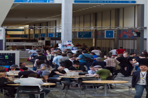 Ein belebter Flughafenvorplatz mit wartenden Menschen, einige stehen und andere arbeiten, Richtungsschilder an den Wänden und an der Decke sowie Deckenleuchten.