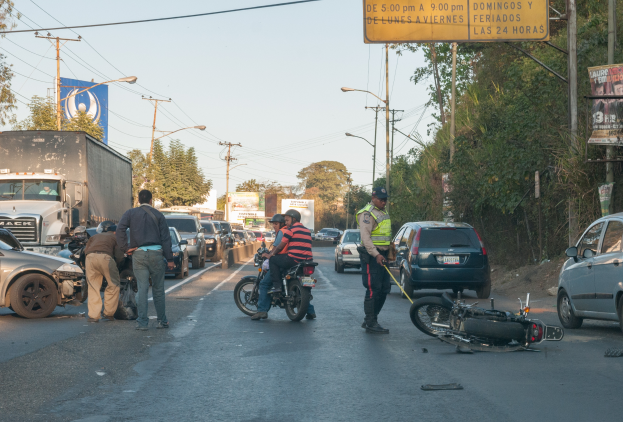 Eine Gruppe von Menschen um ein verunglücktes Motorrad auf der Straße herumsteht, umgeben von mehreren Fahrzeugen, darunter ein Lastwagen, und einem Hintergrund aus Bäumen, Pfosten, Lampen und Schildern unter dem Himmel.