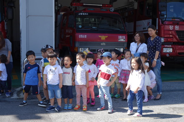 Eine Gruppe von Kindern vor einem Feuerwehrauto an einer Feuerwache stehend, einige mit Mötzen, mit weiteren Feuerwehrautos im Hintergrund.