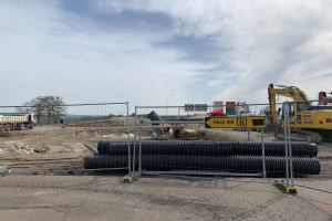 Eine Baustelle mit einem großen Haufen schwarzer Rohre in der Mitte, umgeben von einem Metallzaun, einem Bagger auf der rechten Seite, Fahrzeugen auf der linken Seite und diversen Baustoffen und Bäumen im Hintergrund.