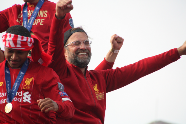 Liverpool-Trainer Jürgen Klopp, der eine rote Jacke trägt, feiert mit seinen Spielern nach dem Gewinn der UEFA Champions League, während im Hintergrund zwei medaillenbekleidete Personen und der Himmel zu sehen sind.