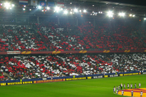 Ein Fußballspiel wird in einem großen Stadion mit Zuschauern auf dem Feld und Sitzplätzen im Hintergrund unter hellen Flutlichtstrahlern gespielt.