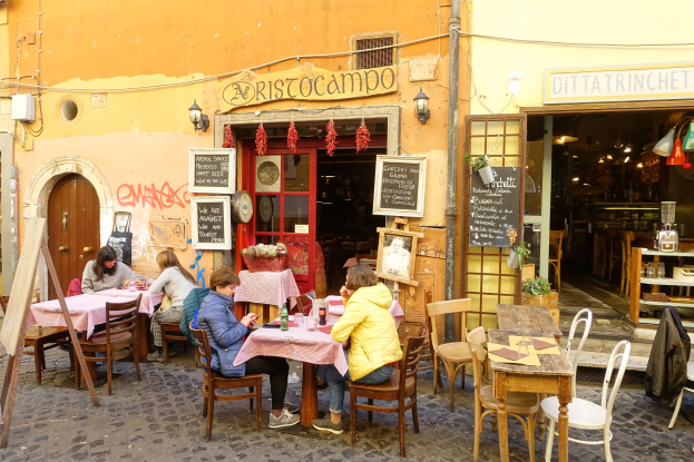 Eine Gruppe von Menschen an Tischen eines Restaurants in Rom, Italien, mit Flaschen, Gläsern und Texttafeln an den Wänden, einer beleuchteten Eingangstür und Regalen mit Gegenständen im Inneren.