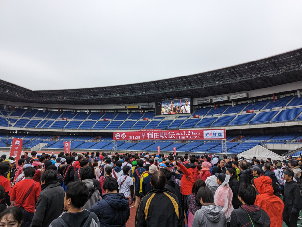 Eine große Menschenmenge steht vor einem Stadion während der Eröffnungszeremonie der Olympischen Spiele Tokio 2020, mit Bannern, Stühlen und einem Bildschirm im Hintergrund unter einem klaren Himmel.
