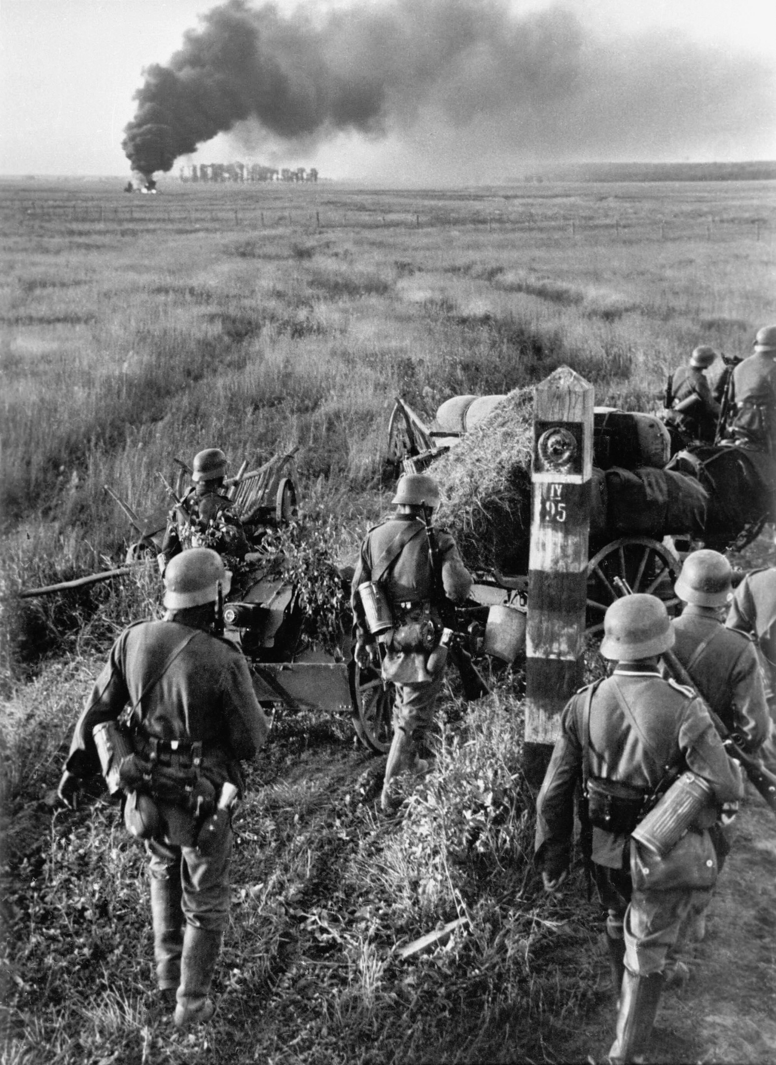 Schwarzes und weißes Bild von deutschen Soldaten in Helmen mit Gewehren, die neben einer Kanone in einem grasbewachsenen Feld mit Rauch im Hintergrund und sichtbarem Himmel stehen.