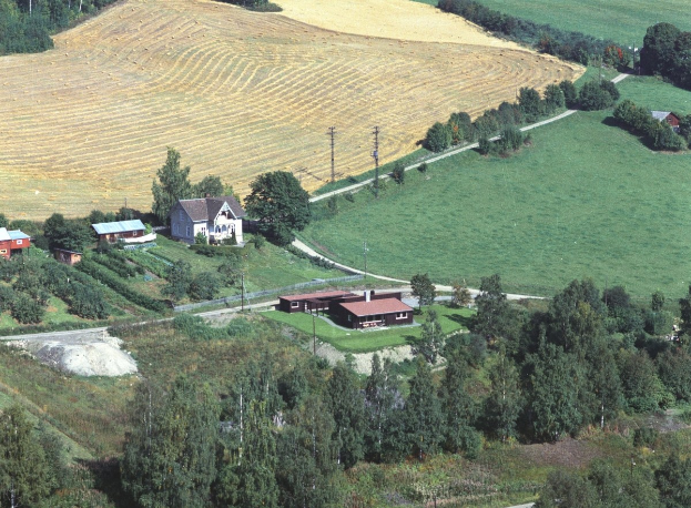 Luftaufnahme eines Bauernhofs in einem grünen Feld umgeben von Bäumen, Häusern, Masten und einer gewundenen Straße.