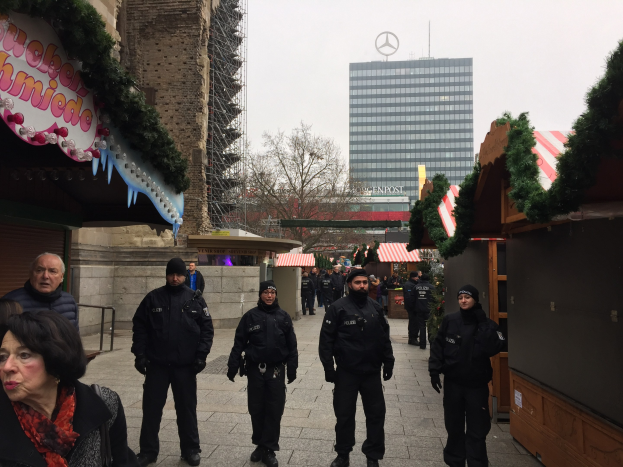 Polizeibeamte in schwarzen Uniformen stehen vor einem geschmützten Weihnachtsmarkt in Berlin, mit Gebäuden, Bäumen und einem klaren Himmel im Hintergrund.