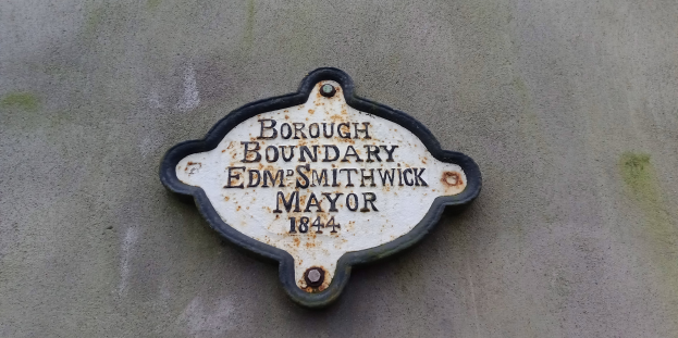 Schwarze Metalltafel an einem Gebäude mit weißer, fetter, zentrierter Schrift, die "Gemeindegrenze Edm Smithwick Bürgermeister" liest.
