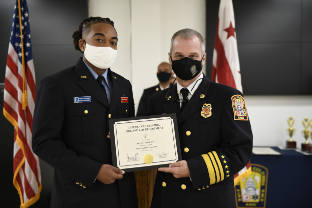 Zwei Männer in Feuerwehrausstattung, einer als Leiter der Feuerwehr des District of Columbia und der andere als stellvertretender Leiter, stehen nebeneinander und tragen Masken sowie ein Zertifikat, mit einem Tisch voller Auszeichnungen und Flaggen im Hintergrund.