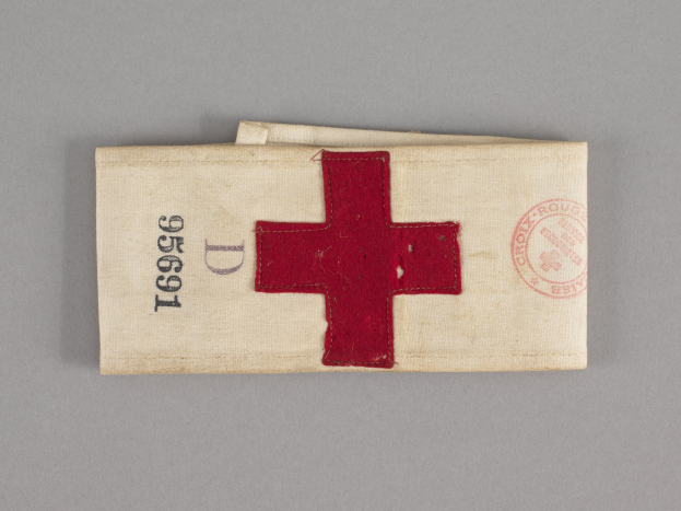 Ein rotes Kreuz-Ersthilfepäckchen in einem Stoffbeutel mit Text und einem Stempel, das auf einer Oberfläche liegt.