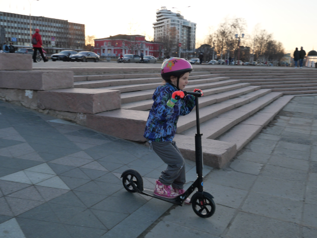 Ein junges Mödchen in einem Helm fährt auf einem Scooter eine Treppe hinunter mit Fahrzeugen, Menschen, Laternen, Bäumen, Gebäuden und einem klaren blauen Himmel im Hintergrund.