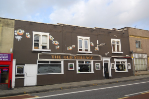 Eine Straßenszene außerhalb der Stadt mit einer Straße, einem Gehweg und Gebäuden mit sichtbaren Türen und Fenstern, darunter eines mit der Aufschrift "Der alte Bottich"; eine rote Wand und eine Glastür auf der linken Seite, ein purpurfarbenes Schild mit Text und ein klarer Himmel darüber.