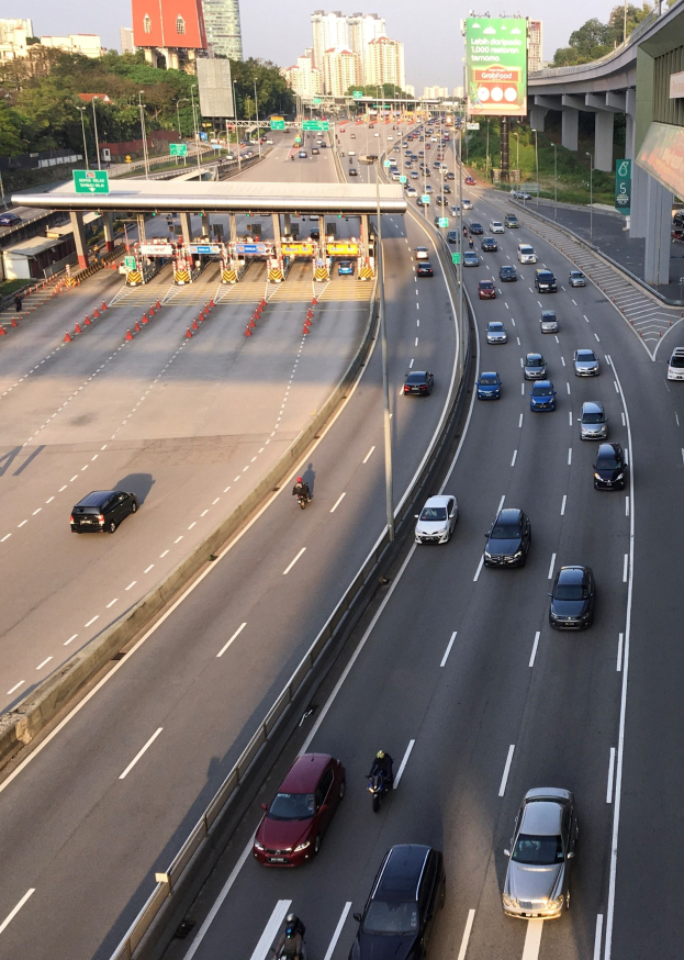 Eine belebte Autobahn mit Autos, Motorrädern, Verkehrsleitkegeln, Straßenschildern, Laternen, einer Brücke, Bäumen, Gebäuden und einem klaren blauen Himmel.