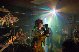 Eine Frau in einer schwarzen Jacke singt in ein Mikrofon auf der Bühne, mit einem Mann in einer Sonnenbrille und einem Stirnband, der ein Instrument spielt, und einem 'LA Guns'-Plakat und Diskoleuchten im Hintergrund.