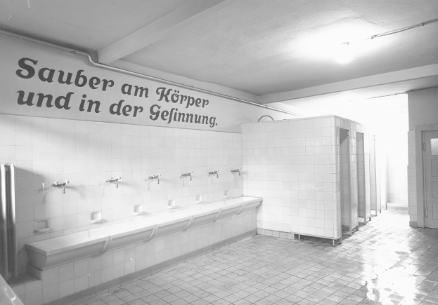 Schwarz-weiß-Foto einer öffentlichen Toilette mit Fliesenboden, einer textbedeckten Wand, mehreren Wasserhähnen, einer Tür und einer deckenmontierten Lampe.