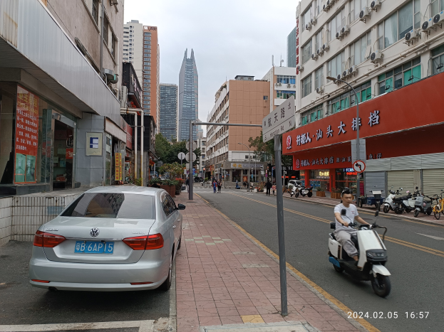 Ein Mann fährt mit einem Roller die Straße einer Stadt entlang, die von hohen Gebäuden gesäumt ist, mit Fußgängern auf den Bürgersteigen und anderen Fahrzeugen auf der Straße, unter einem klaren blauen Himmel mit Bäumen im Hintergrund.