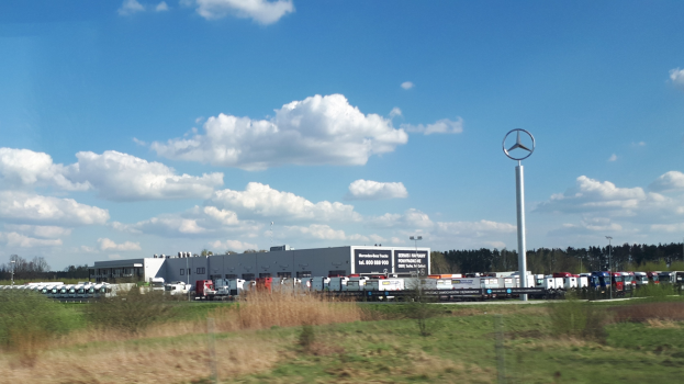Mercedes-Benz-Fabrik in Deutschland mit Fahrzeugen auf der Straße, umgeben von Grünflächen und Gebäuden, unter einem bewölkten Himmel.