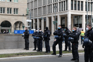 Eine Gruppe von Polizisten in schwarzen Uniformen und Masken steht vor einem gläsernen Gebäude mit Säulen in Berlin, Deutschland, einige halten Helme, mit Laternenmasten, Verkehrsampeln, Texttafeln, einer Statue, anderen Menschen und grasbewachsenem Boden im Hintergrund.